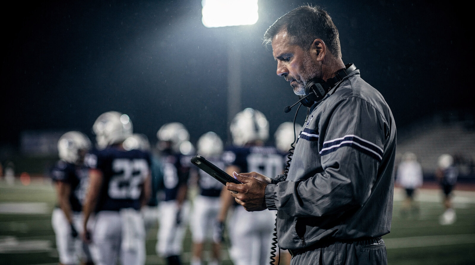 Coach in a headset checking game stats on a tablet on the sideline under the lights