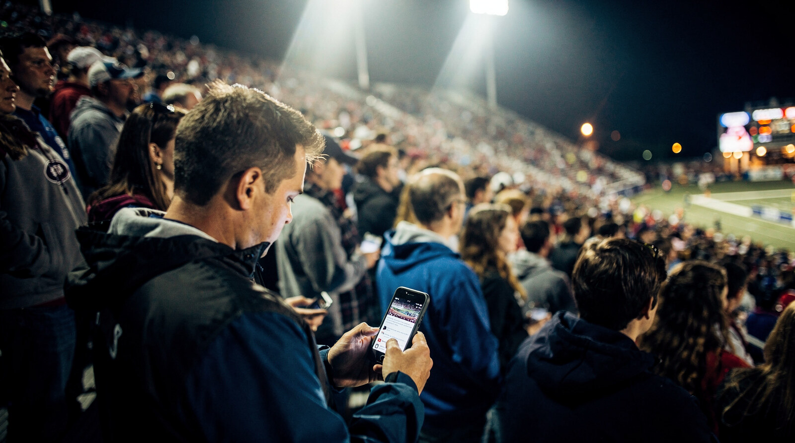 Fan in a packed night crowd following the live game feed on a phone